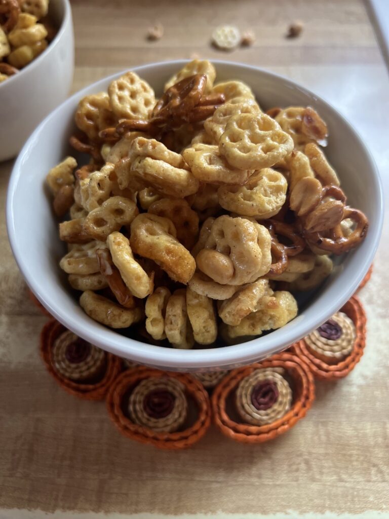 A white bowl filled with Honey Cereal Snack Mix, featuring golden cereal pieces, pretzels, peanuts, and honey styled on a kitchen countertop.