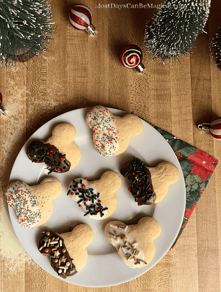 Overhead view of Mickey-shaped Christmas dipped sprinkle cookies on a white plate with a poinsettia napkin, ornaments, and small Christmas tree decorations in the background.