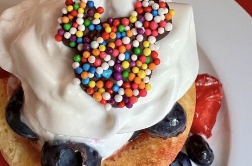Chocolate decorations shaped like everyone's favorite mouse with colorful nonpareils on a strawberry and blueberry shortcake, served on a white plate.