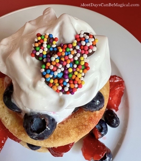 Chocolate decorations shaped like everyone's favorite mouse with colorful nonpareils on a strawberry and blueberry shortcake, served on a white plate.