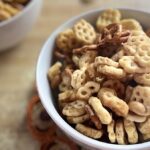 A white bowl filled with Honey Cereal Snack Mix, featuring golden cereal pieces, pretzels, peanuts, and honey styled on a kitchen countertop.