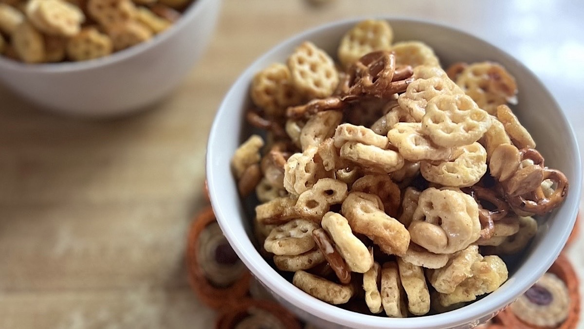 A white bowl filled with Honey Cereal Snack Mix, featuring golden cereal pieces, pretzels, peanuts, and honey styled on a kitchen countertop.
