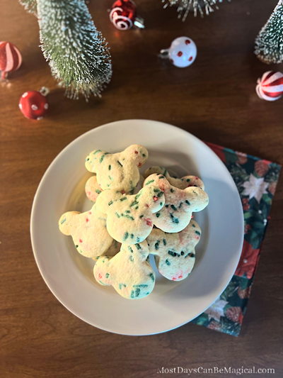 A plate of homemade Mickey-shaped Christmas Confetti Cookies with red & green sprinkles, set against a background of festive holiday decorations.