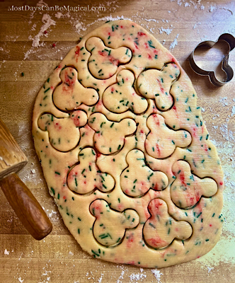 Rolled out Christmas Confetti Cookie dough with Mickey shapes cut out but still inside the dough, a Mickey-shaped cookie cutter in the corner, a rolling pin peeking in on the side, and scattered sprinkles and flour on the surface underneath.