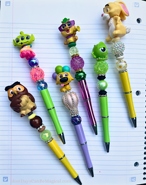 Six Disney-inspired handmade pens are arranged on a piece of notebook paper. Each pen features a different character and color scheme keeping whimsy in the forefront.