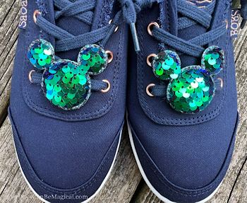 A pair of Mickey shoe clips decorated with blue and green sequins clip to a pair of navy lace-up shoes.