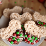 Christmas Mickey-shaped Rice Krispie treats, partially dipped in chocolate with white chocolate drizzle and red and green M&Ms, arranged on a white plate with scattered M&Ms and Christmas decorations in the background.