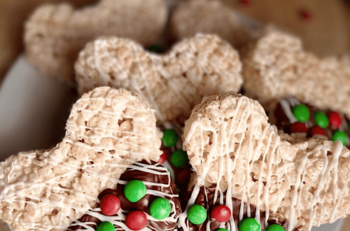 Christmas Mickey-shaped Rice Krispie treats, partially dipped in chocolate with white chocolate drizzle and red and green M&Ms, arranged on a white plate with scattered M&Ms and Christmas decorations in the background.