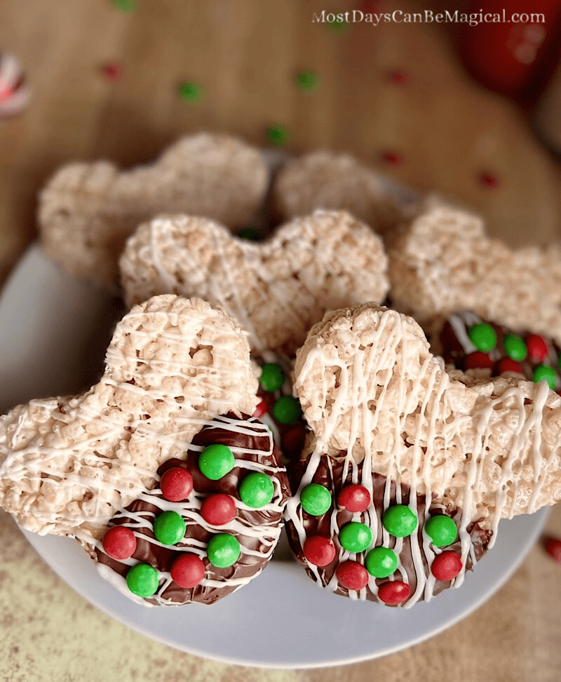 Christmas Mickey-shaped Rice Krispie treats, partially dipped in chocolate with white chocolate drizzle and red and green M&Ms, arranged on a white plate with scattered M&Ms and Christmas decorations in the background.