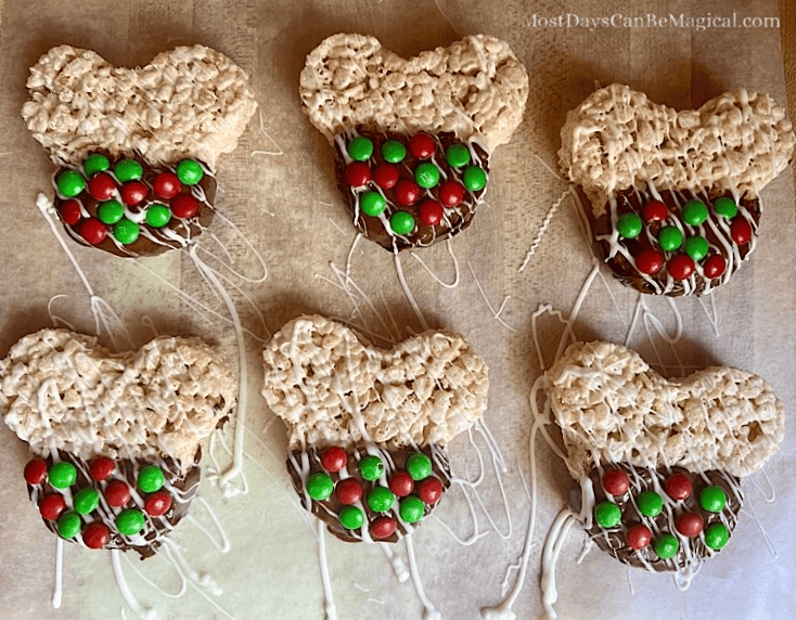 Freshly dipped Mickey-shaped Rice Krispie treats with white drizzle and red and green M&Ms, drying on wax paper.