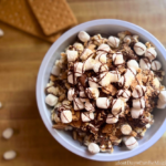 Overhead view of a s'mores popcorn bowl filled with popped corn, drizzled chocolate, mini marshmallows, crushed graham crackers, and toasted marshmallows on top. A cozy, indulgent treat inspired by Disney-style snacks.