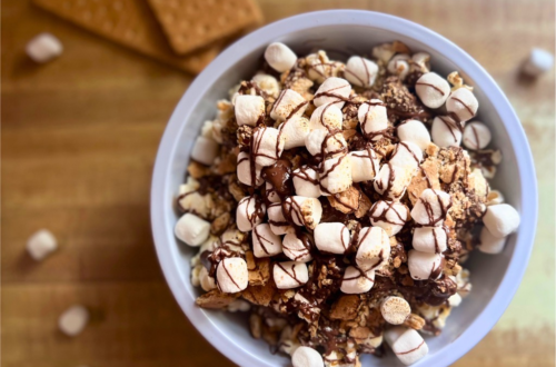 Overhead view of a s'mores popcorn bowl filled with popped corn, drizzled chocolate, mini marshmallows, crushed graham crackers, and toasted marshmallows on top. A cozy, indulgent treat inspired by Disney-style snacks.