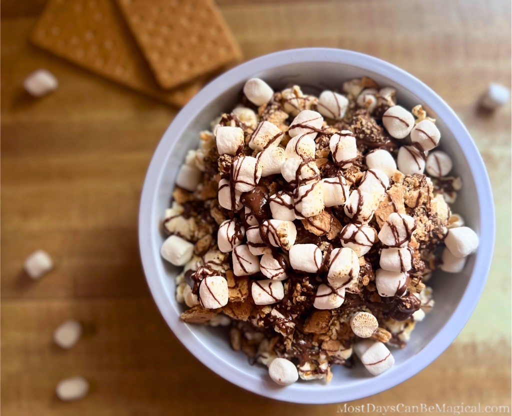 Overhead view of a s'mores popcorn bowl filled with popped corn, drizzled chocolate, mini marshmallows, crushed graham crackers, and toasted marshmallows on top. A cozy, indulgent treat inspired by Disney-style snacks.