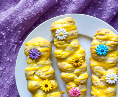 Three golden sweet braids drizzled with lemon glaze and decorated with colorful edible flowers on a white plate over a purple tablecloth.