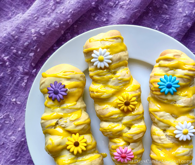 Three golden sweet braids drizzled with lemon glaze and decorated with colorful edible flowers on a white plate over a purple tablecloth.
