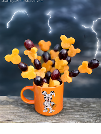 Cup of Disney-inspired Halloween Fruit skewers featuring Mickey-shaped cantaloupe and grapes with a stormy sky background.