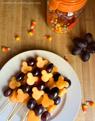 Disney-inspired fruit skewers featuring Mickey-shaped cantaloupe and grapes are resting on a plate surrounded by candy corn and grapes.
