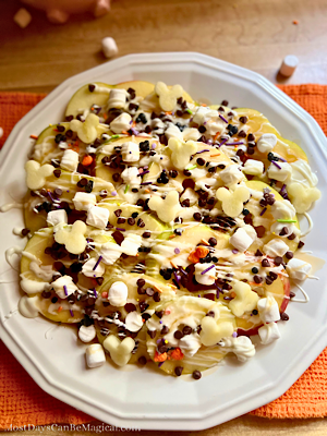 Overhead view of Disney-inspired Caramel apple nachos on a white plate topped with mini marshmallows, festive sprinkles, caramel, white chocolate, mini chocolate chips, and sea salt.