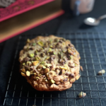 Colossal Pistachio Toffee Cookie on a cooling rack with a large pink book and small mug with spoon on the dark background and a few pistachios sprinkled around.