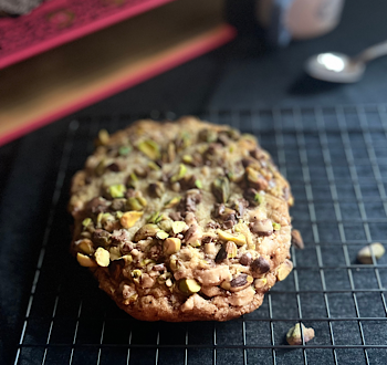 Colossal Pistachio Toffee Cookie on a cooling rack with a large pink book and small mug with spoon on the dark background and a few pistachios sprinkled around.