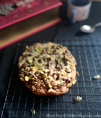 Colossal Pistachio Toffee Cookie on a cooling rack with a large pink book and small mug with spoon on the dark background and a few pistachios sprinkled around.